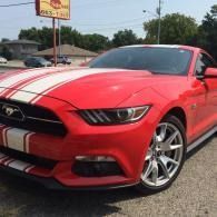 Red Ford Mustang with white racing stripes parked outdoors on a sunny street.