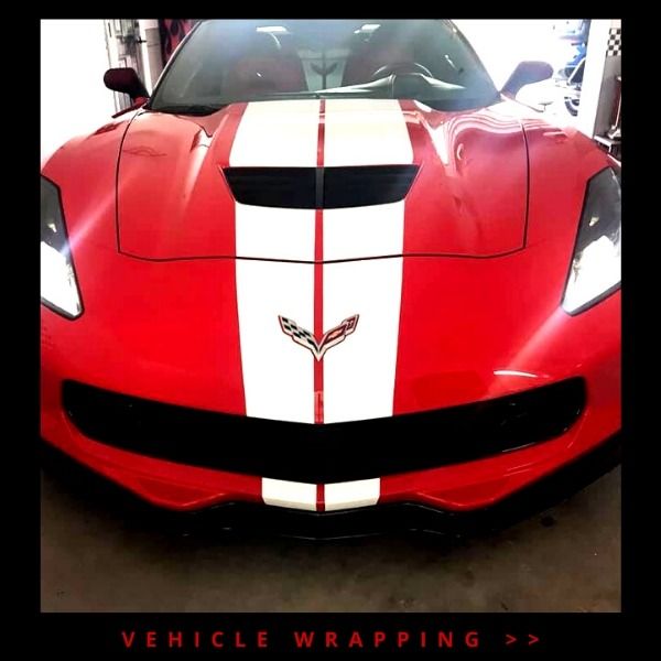 Red Chevrolet Corvette with white racing stripe and black accents in a vehicle wrap shop