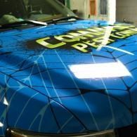 Blue car hood with black web-like lines and bold yellow lettering under bright indoor light