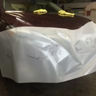 Car covered with a white protective sheet in a garage, maroon roof visible, yellow cloths on top