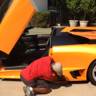Man kneeling beside an orange sports car with its door raised, inspecting the front wheel area outdoors.