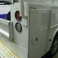 White utility truck side with rear lights and a small access door, parked on a paved surface