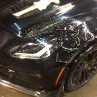 Black sports car front corner with a bright headlight and wet, reflective paint under indoor lights