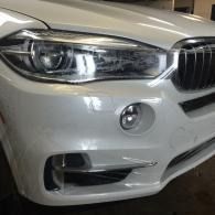 Front of a white BMW sedan showing the headlight, grille, and fog light in a garage.