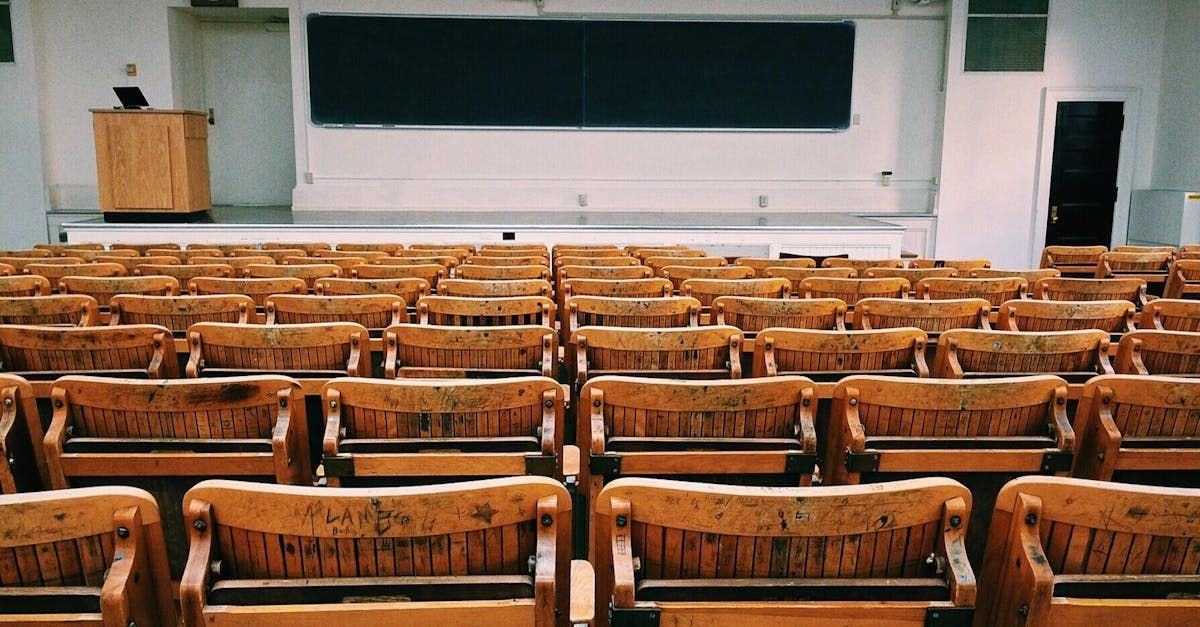 Rows of wooden chairs in an auditorium with a blackboard in the background.