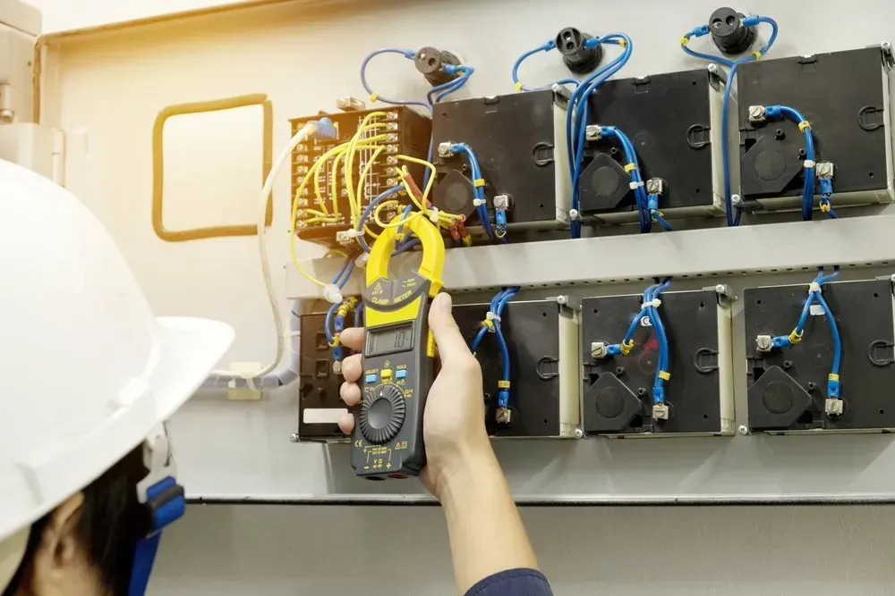 Electrician in a hard hat uses a clamp meter to inspect electrical panel wiring.