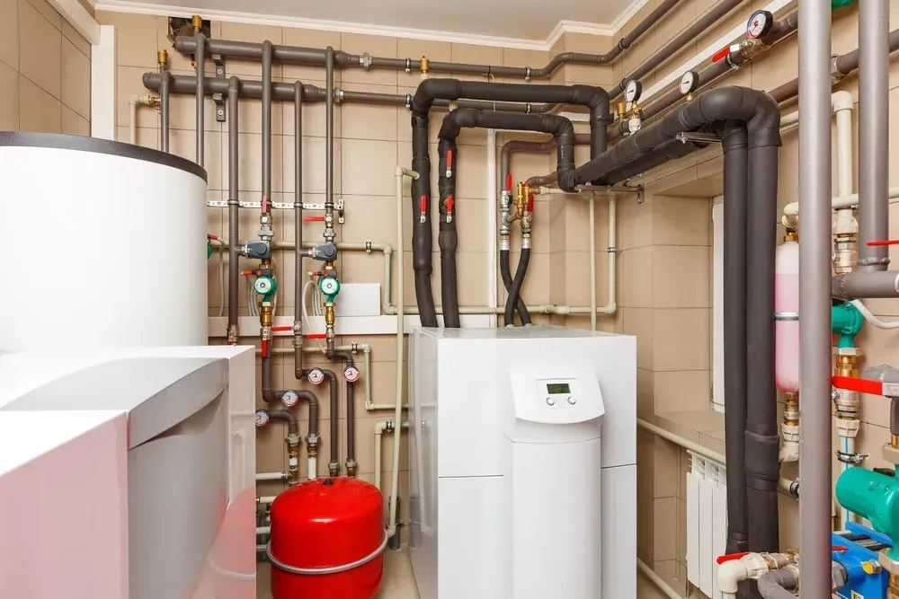 Plumbing and heating equipment in a utility room with pipes, tanks, and gauges; mostly neutral colors.