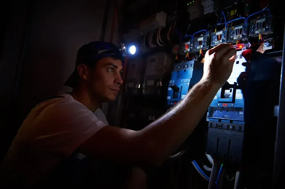 A man in a cap using a flashlight to work on an electrical panel in a dim room.