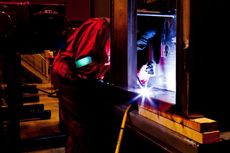 Welder in red protective gear, working on a metal surface. Bright arc visible.