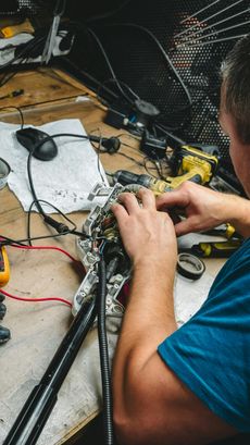 A person with hands on a circuit board with wires, tools, and a multimeter on a messy workbench.