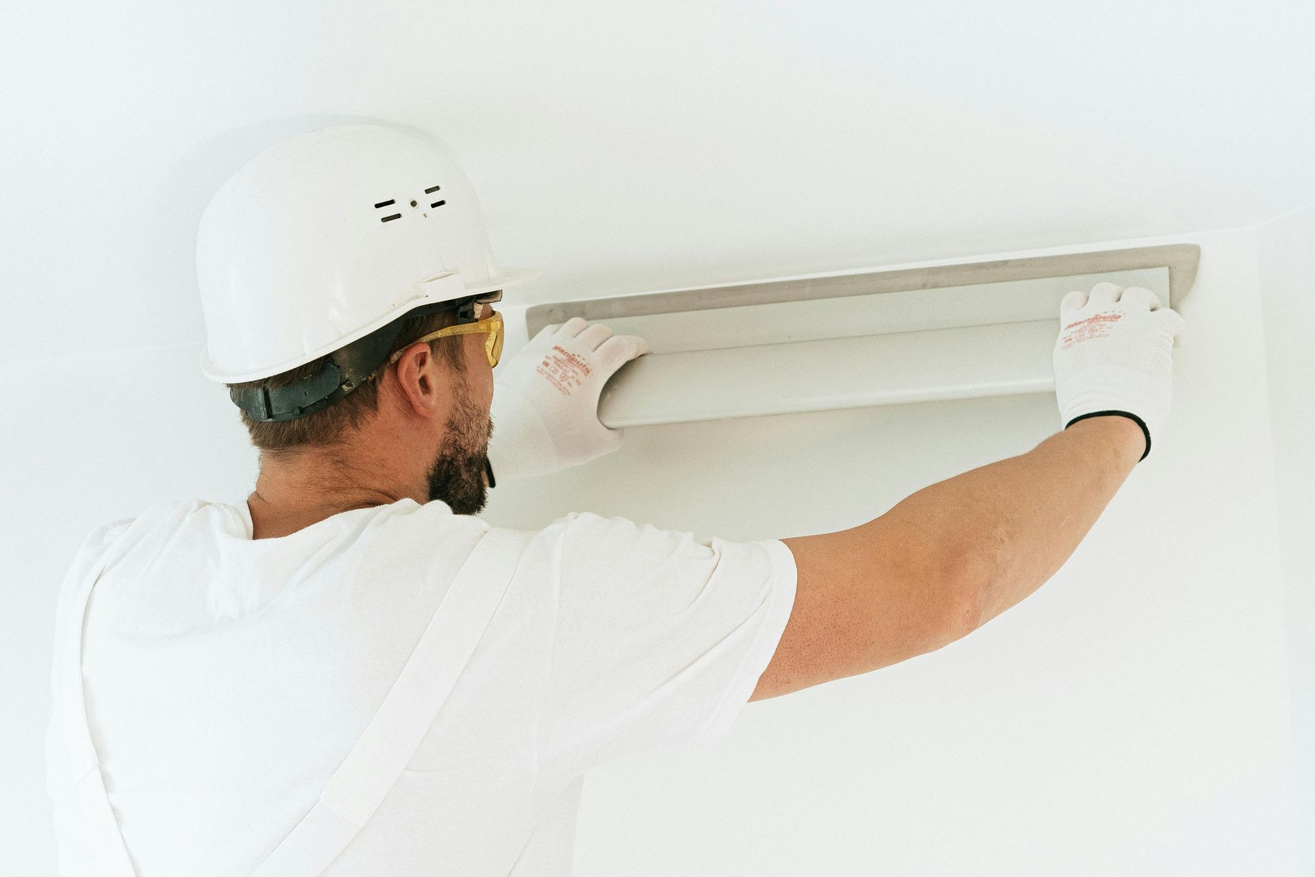 Construction worker in white, installing a rectangular air duct; wearing gloves, goggles, and helmet.