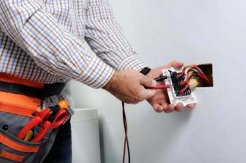 Electrician testing wires in a wall outlet; wearing work belt, indoors.
