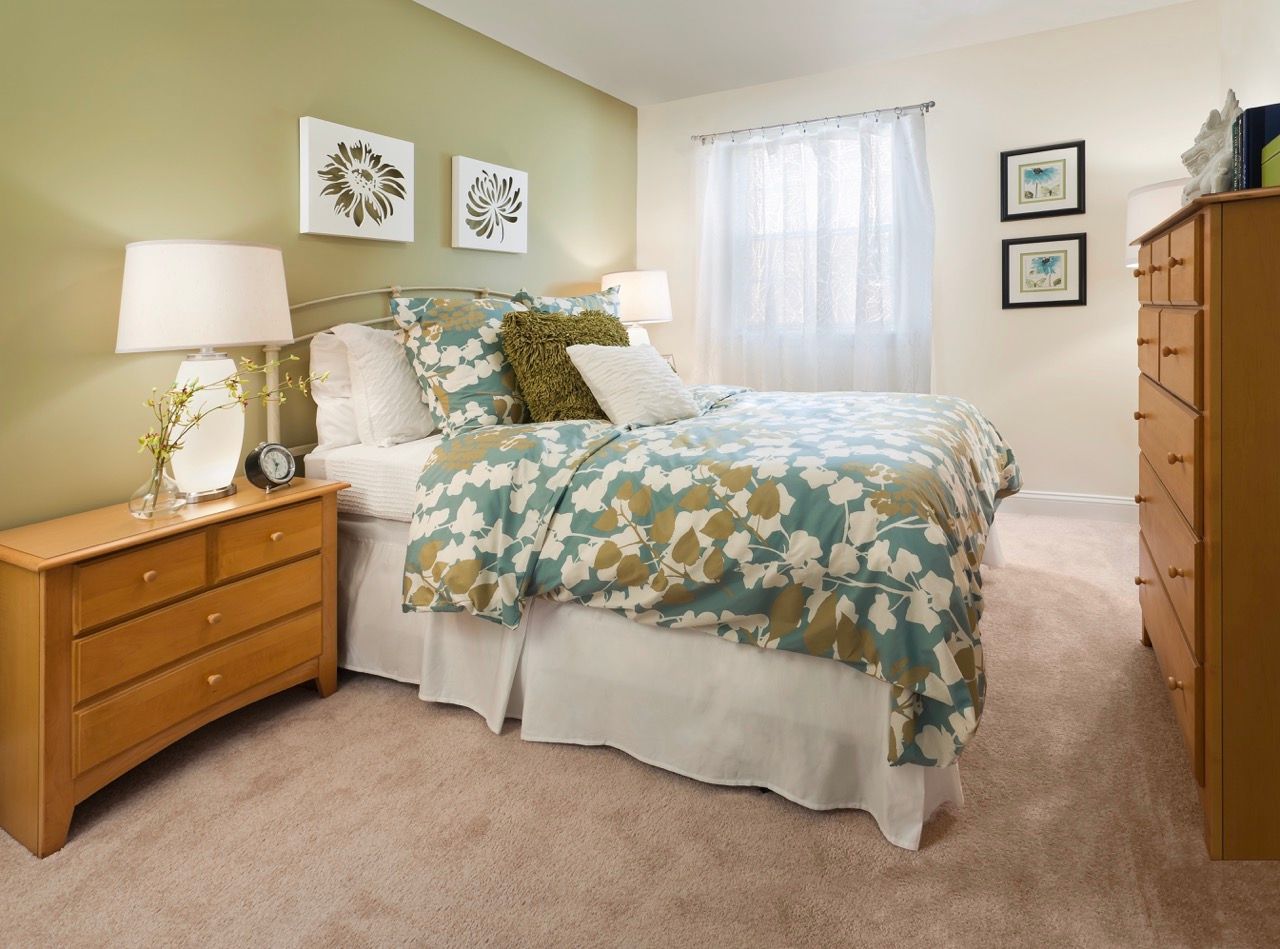 Interior bedroom in an apartment with a queen bed, teal and tan floral bedding, two nightstands, and a window with sheer curtains.