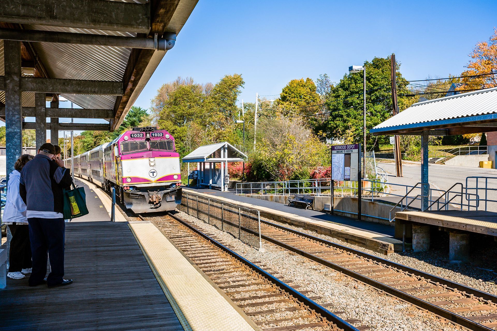 Pink train approaching a sunlit station platform with people waiting nearby