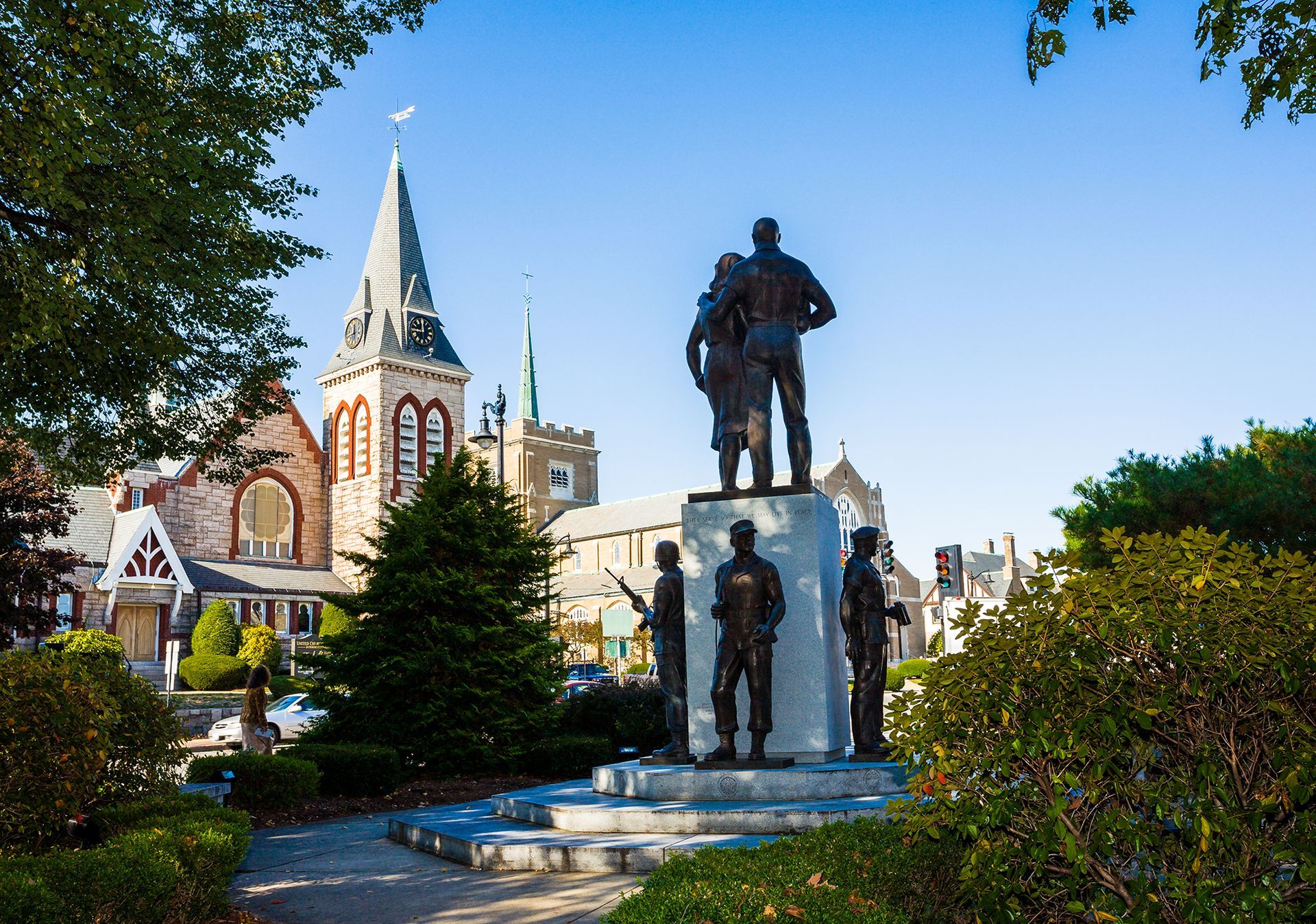 Statue in a town square beside a church, with green trees and a clear blue sky.