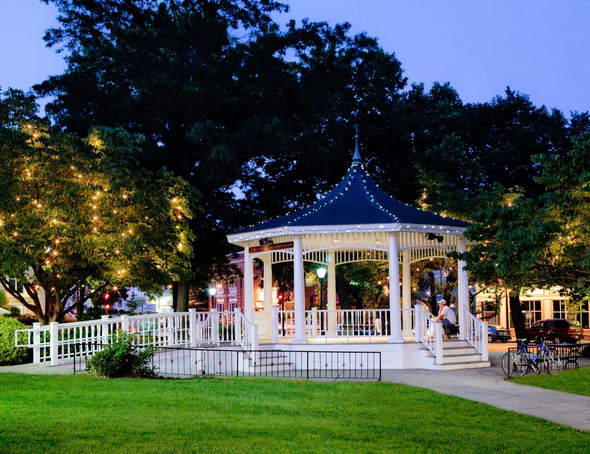 White gazebo lit at dusk in a park, surrounded by trees, grass, and a picket fence
