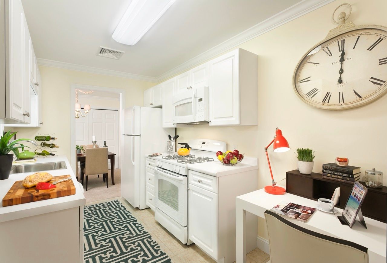 Bright white apartment kitchen with stove, fridge, and a small desk along the wall.