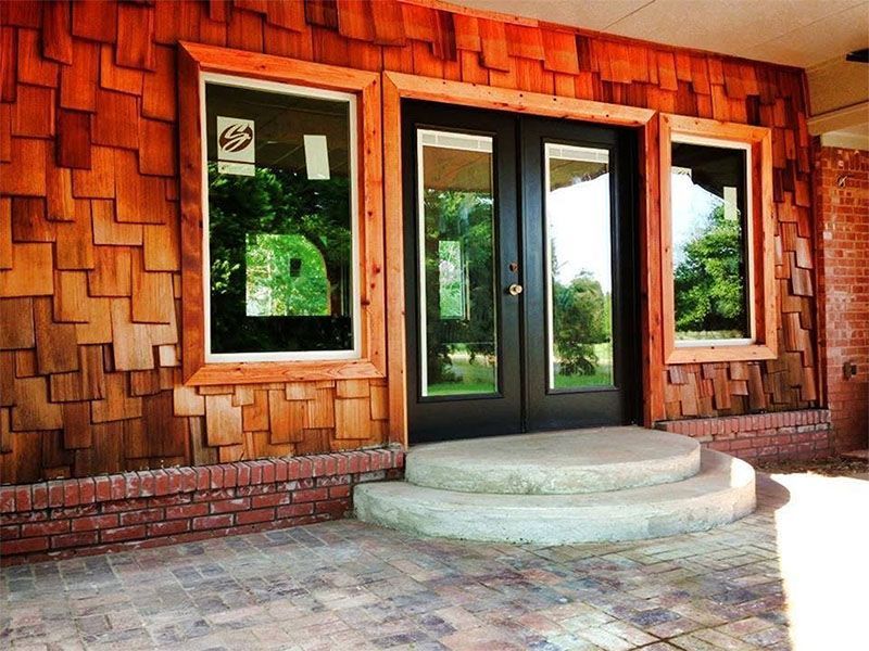 Wooden-shingled building facade with black double doors, windows, and brickwork; concrete steps.