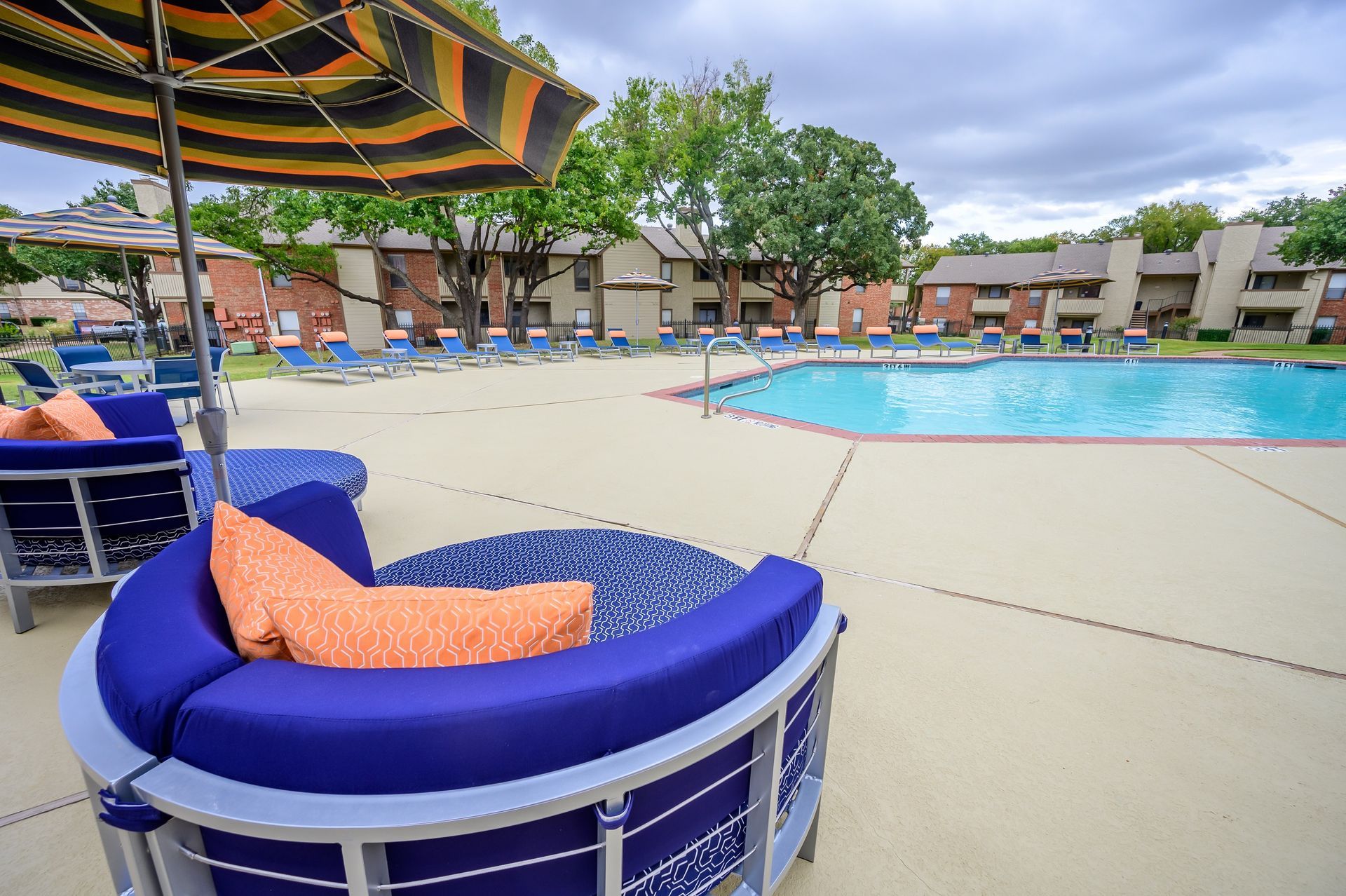 Poolside lounge area with blue seating, orange cushions, umbrella, and apartment buildings in the background.