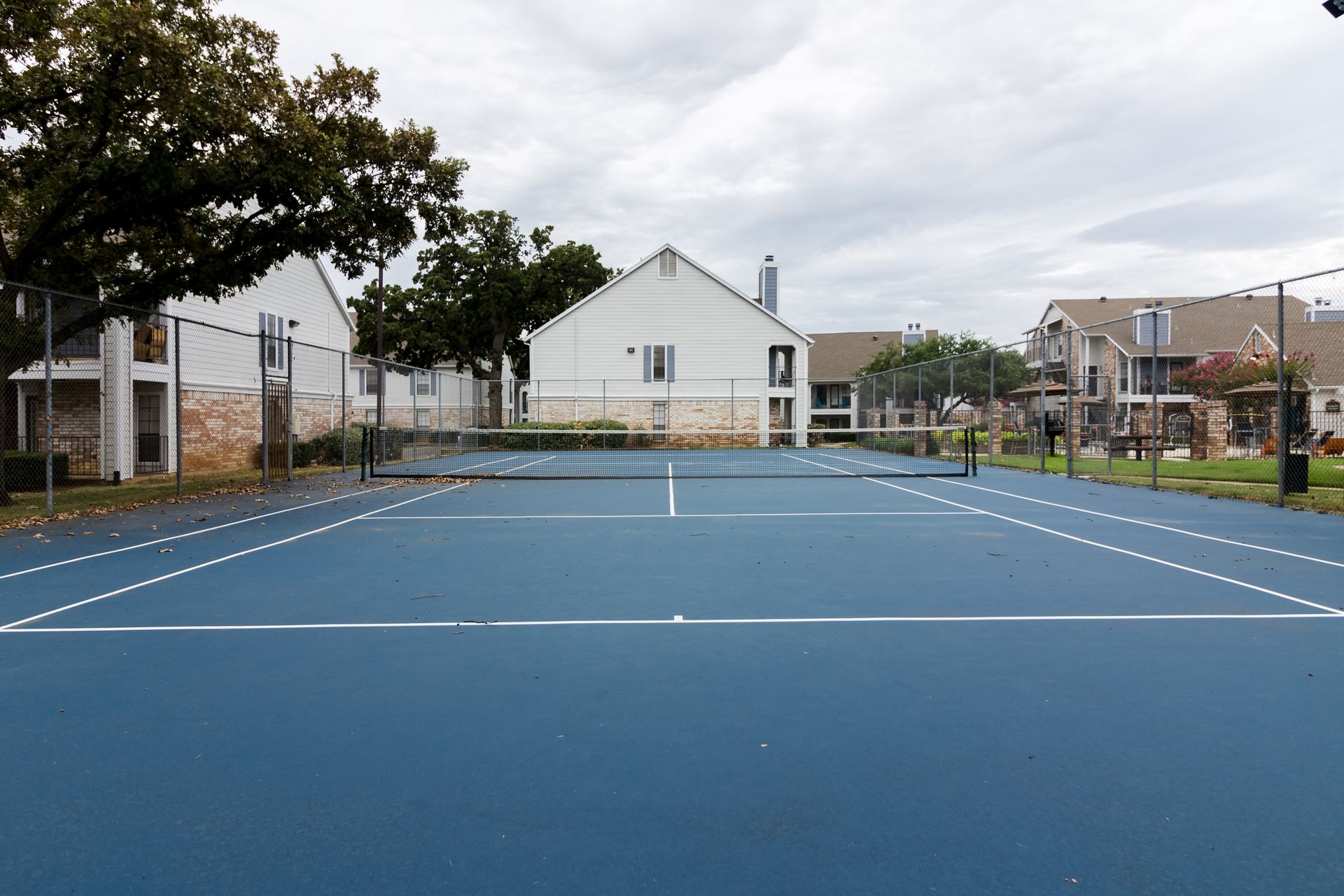 Blue tennis court, white lines, net. Buildings in background, overcast sky.