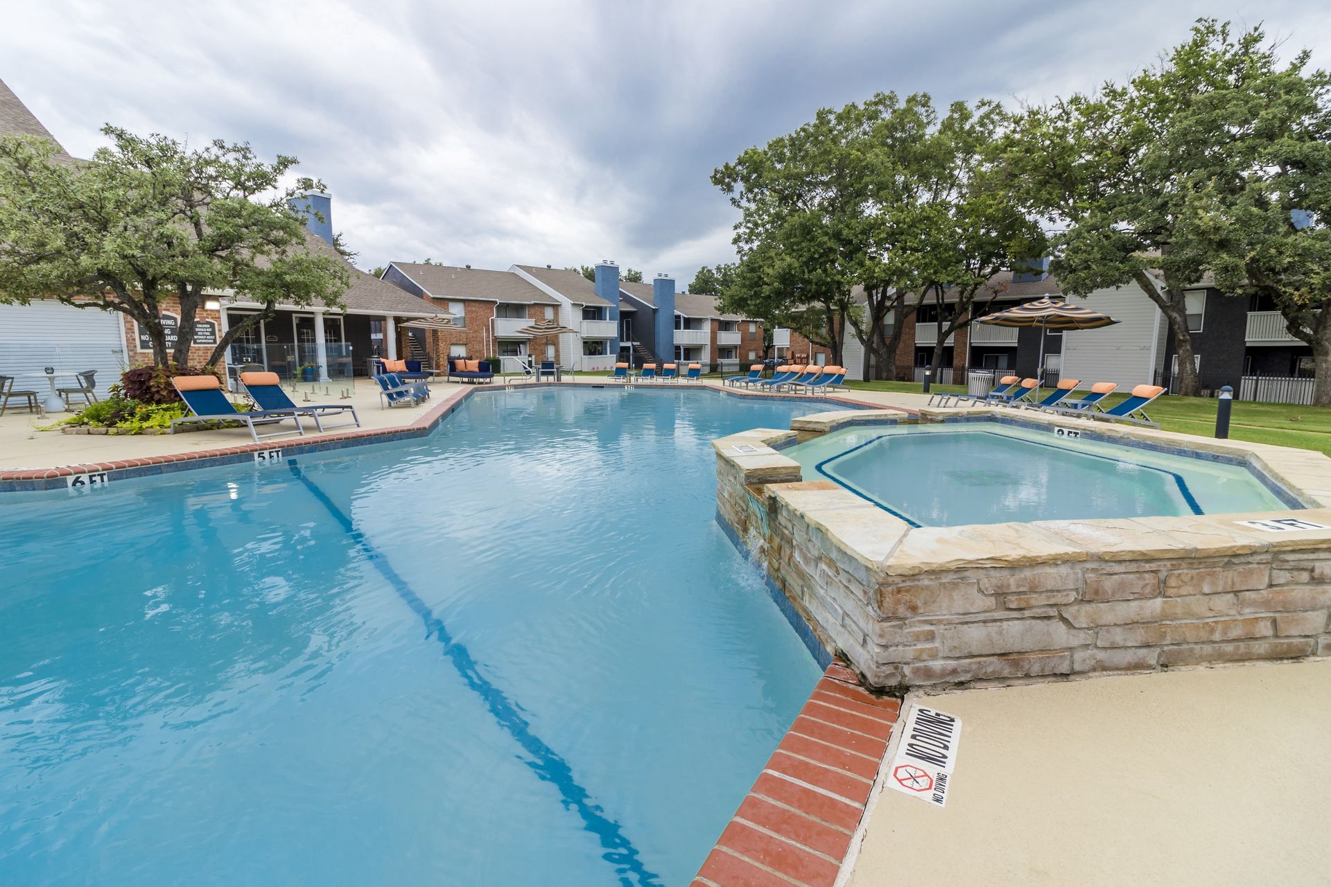 Swimming pool and hot tub at an apartment complex on an overcast day, surrounded by trees and lounge chairs.