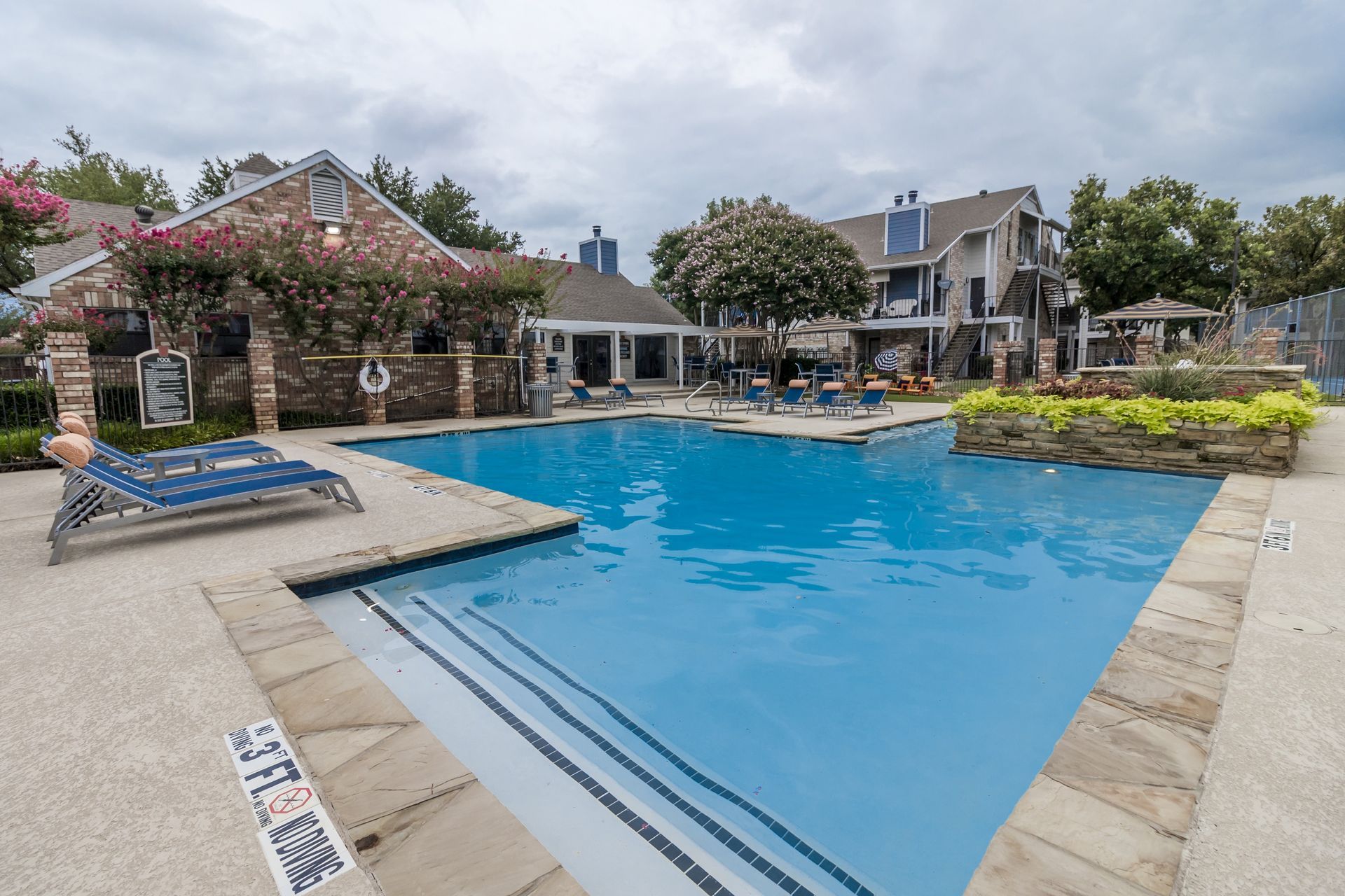 Swimming pool in front of an apartment complex with cloudy skies. A person lounges on a chair.