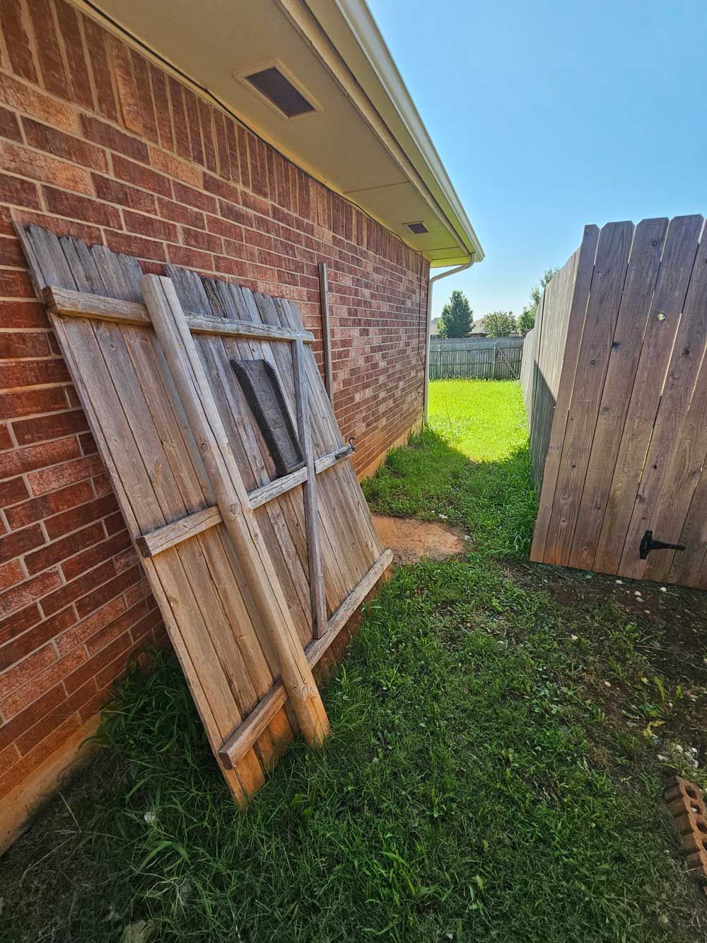 A leaning wooden fence panel next to a brick wall, next to a narrow grassy pathway.