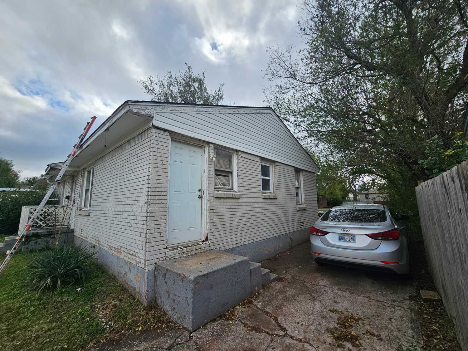 Small, white-sided house with a door, a car parked in the driveway, and a ladder leaning on the roof.