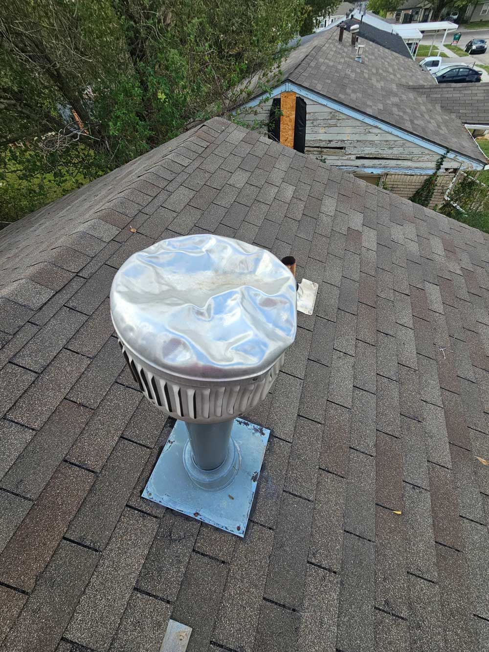 A distorted metallic roof vent on a brown shingled roof, with other buildings visible in the background.