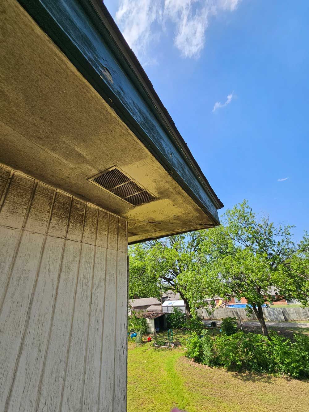 Corner of a building with a square vent. Light blue sky and green trees are in the background.