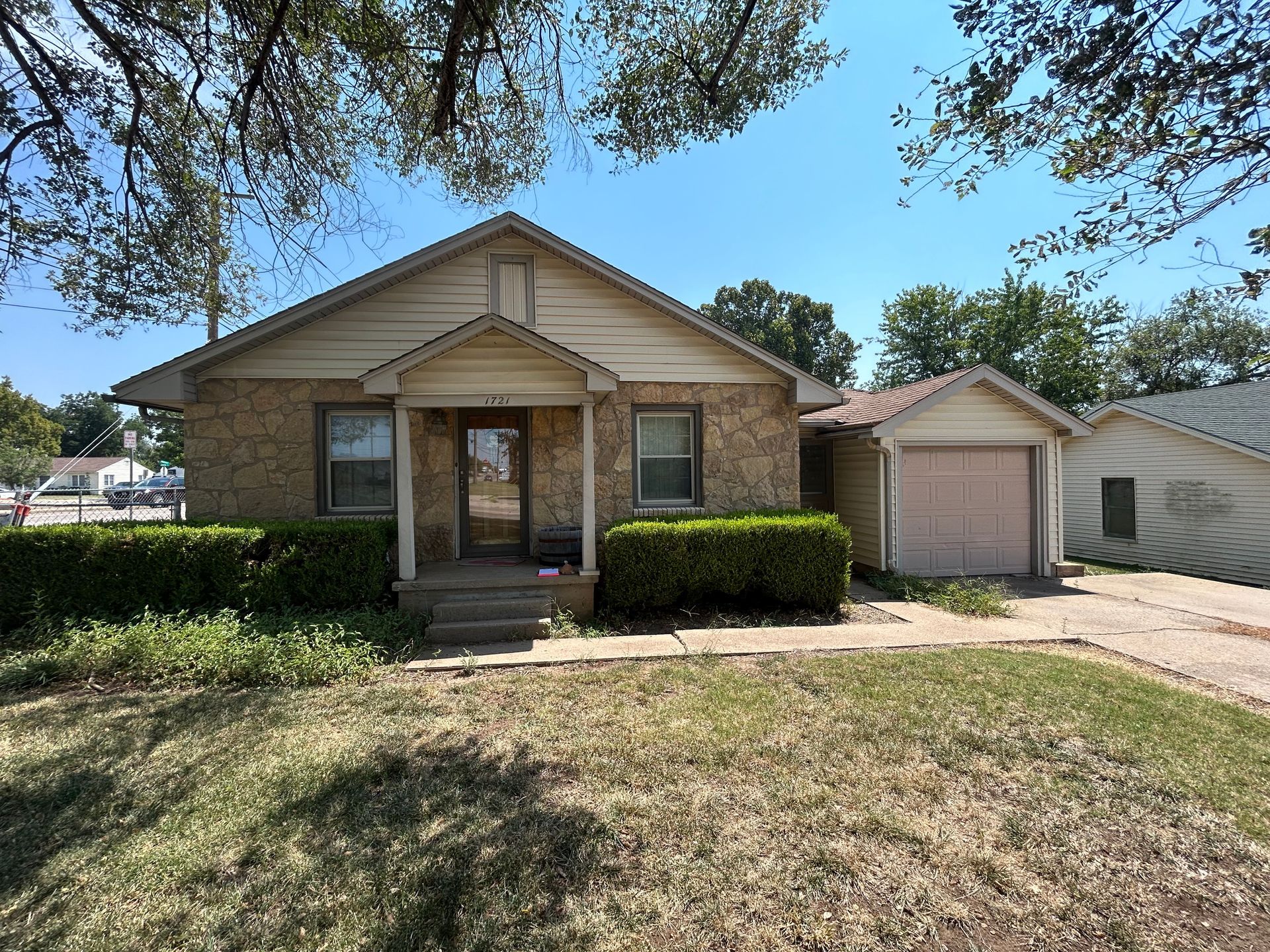 Small, brick house with green bushes, driveway, and attached garage on a sunny day.