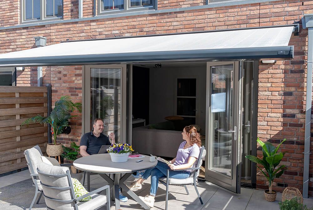 Een man en een vrouw zitten aan een tafel op een terras voor een bakstenen gebouw.