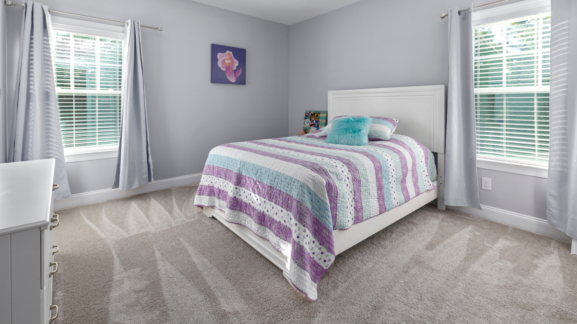 Bedroom with a white bed, patterned quilt, two windows with gray curtains, gray walls, and light carpet.