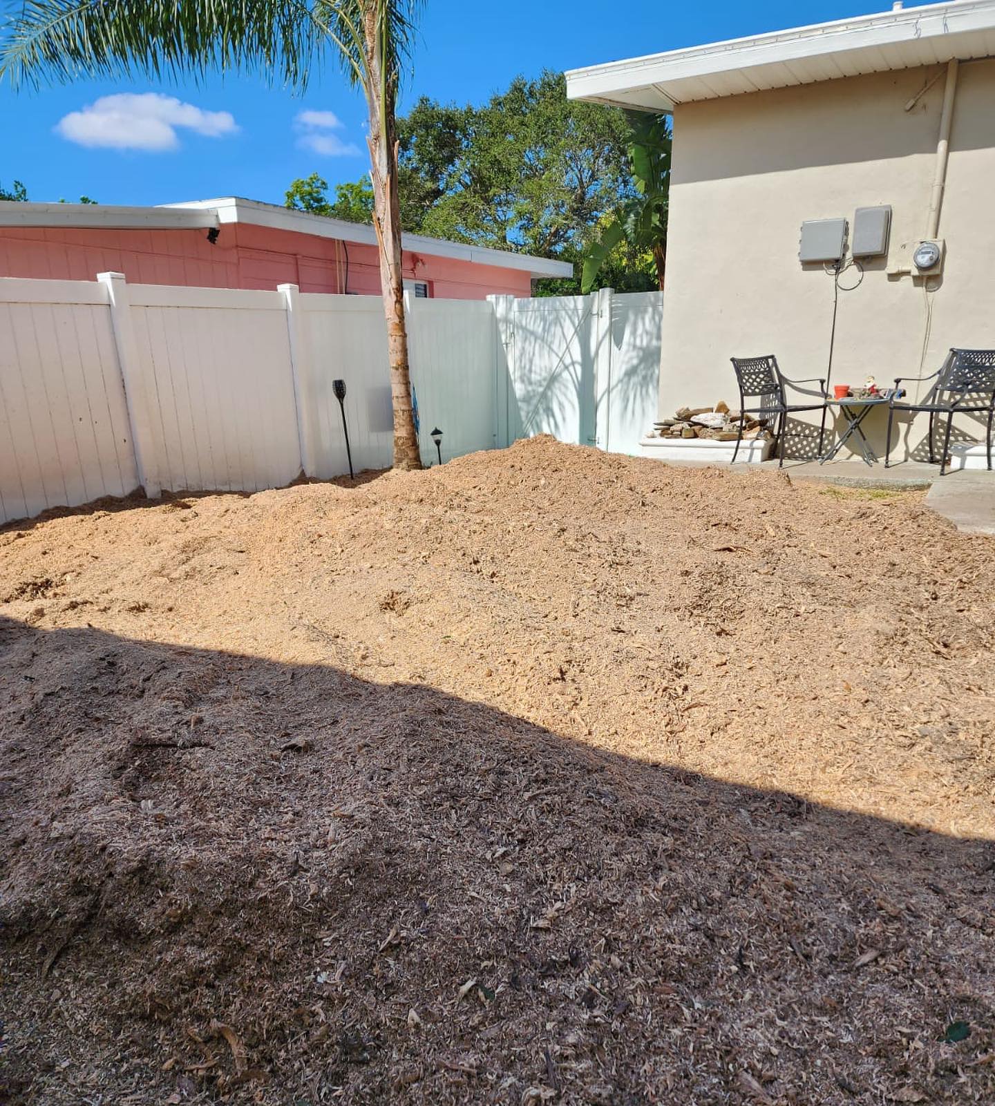 A large pile of wood chips covers a backyard, with a white fence and a building.