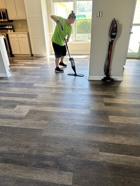 Person mopping a floor with a spray mop in a room with wood-look flooring. A vacuum cleaner is nearby.