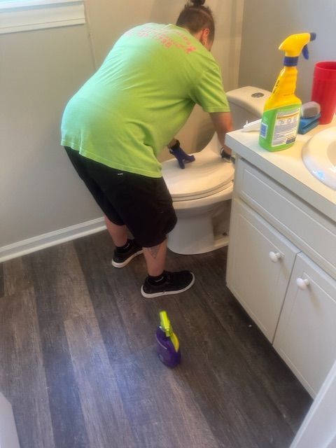Person cleaning a toilet in a bathroom; wearing a green shirt and black shorts; cleaning supplies visible.