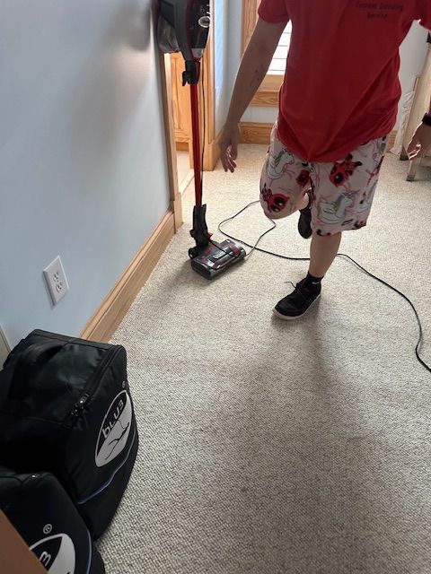 Person in shorts vacuums a carpeted hallway; vacuum cleaner is upright.
