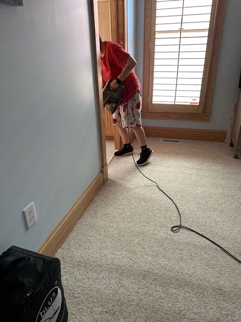 Person in red shirt and shorts using a vacuum in a room with carpet and a blue wall.