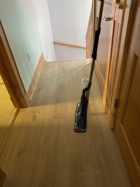 A person is cleaning a light-colored wooden floor with a mop in a hallway, stairs in the background.