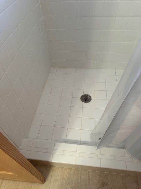 White tiled shower with drain, partially obscured by a shower curtain, wood floor visible below.