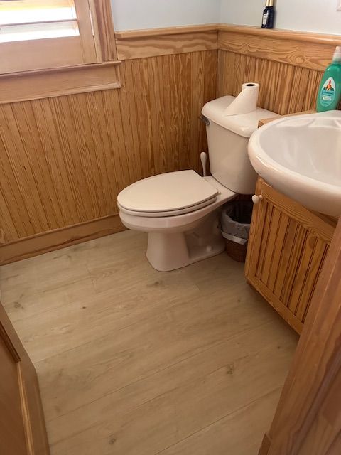 Small bathroom with wood paneling, a toilet, sink, and light wood-colored floor.