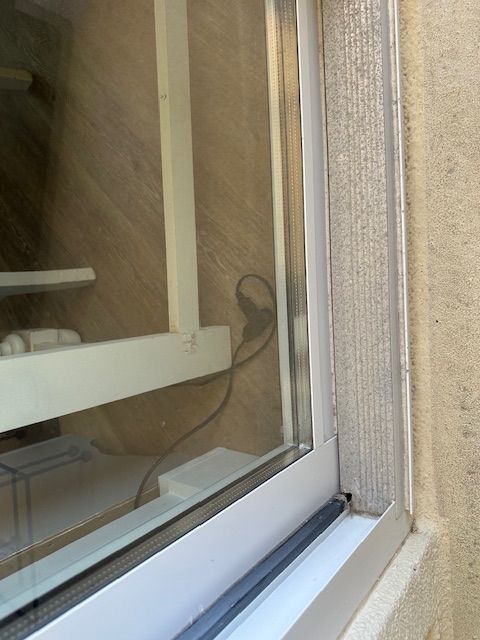 Close-up of a window frame with a light-colored wall, a white desk, and a power cord visible through the glass.