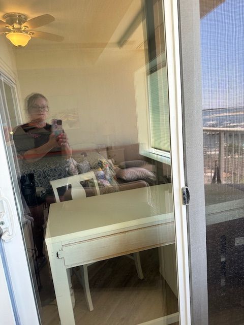Person reflected in a sliding glass door taking a selfie. Interior space with a white table, sofa, and window.
