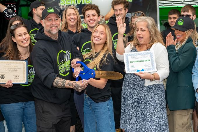 Group holding a blue ribbon and awards, smiling in front of a green sign outdoors. | East End Auto Kicks