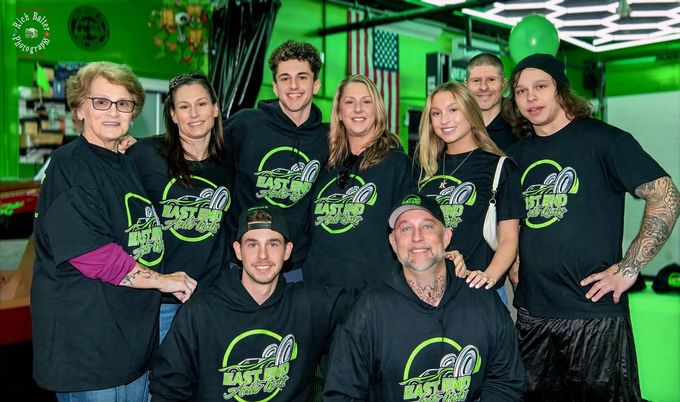 Group of nine people in matching black shirts posing indoors against a green-lit background | East End Auto Kicks