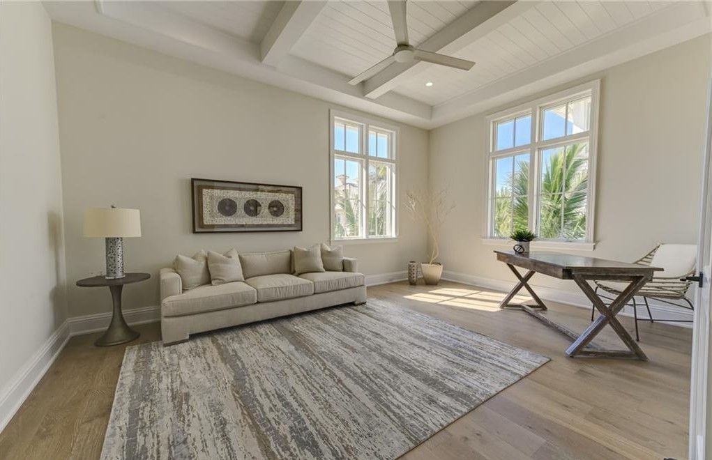 Room with sofa, rug, desk, two windows, and light wood flooring. Neutral color palette with light beige walls.