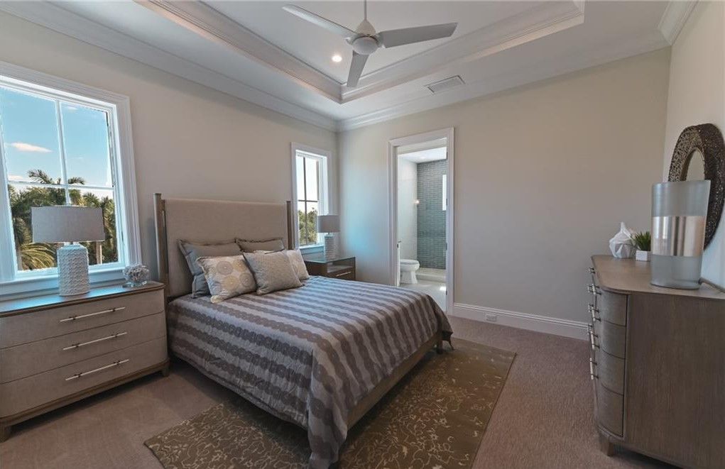 Bedroom with gray and beige tones, large window, bed, dresser, ceiling fan.