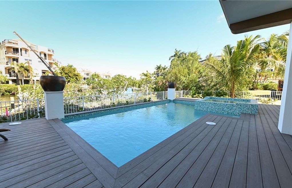 Pool with wooden deck overlooking a body of water, trees, and buildings; blue sky.