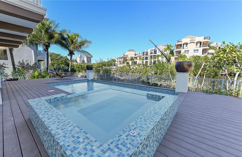 Triangular pool with blue mosaic tile, on wooden deck, sunny day, with palm trees and buildings in background.