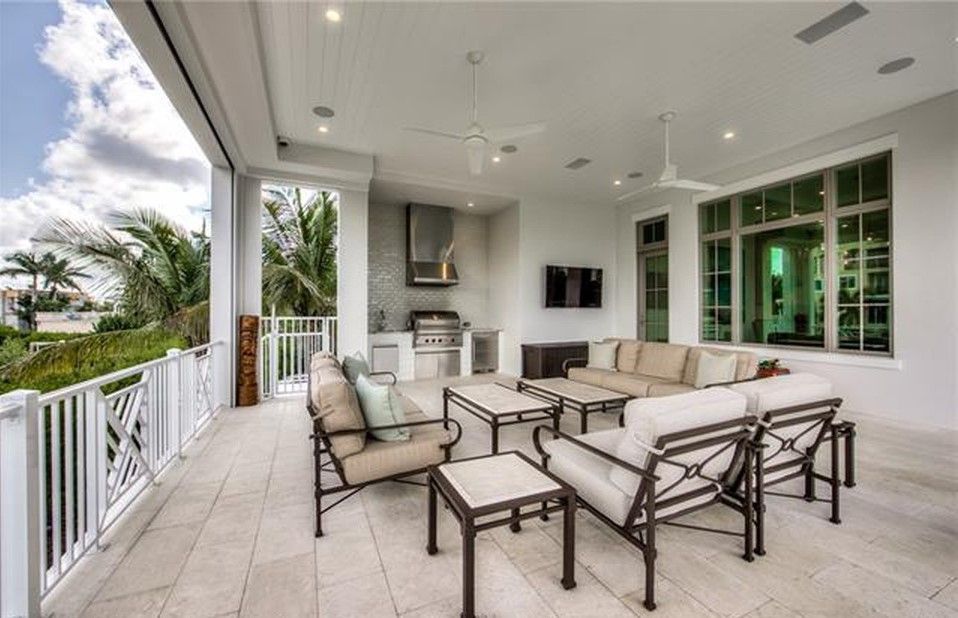 Outdoor patio with seating, grill, TV, and large windows. White walls and railings with blue sky.
